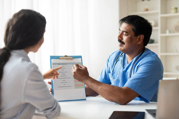 Mature Indian Doctor Consulting Patient Before Medical Check Up In Clinic, Professional Therapist Male Showing Clipboard And Answering Questions About Treatments During Appointment In Office, Closeup