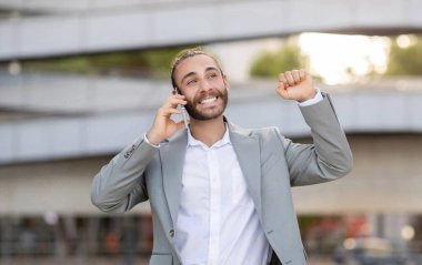 Neşeli genç işadamı açık hava telefon görüşmesi sırasında başarıyı kutluyor, mutlu erkek girişimci iş ortağıyla cep telefonu görüşmesi yapıyor, zafer için elini kaldırıyor, iyi haberler aldı.