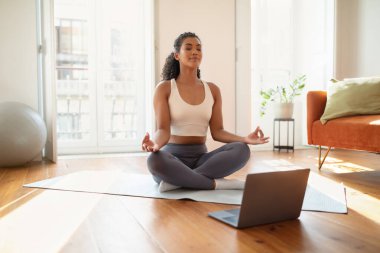Huzurlu genç kadın, rehberli internet yogası sırasında laptopta bilinçlilik meditasyonu yapıyor, sabah evde lotus pozisyonunda oturuyor, dengeli spor yaşam tarzını somutlaştırıyor.