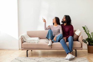 Heyecanlı neşeli genç Hintli kadın ve adam evdeki kanepede oturmuş, uzaktan kumandayı kullanarak soğuk havada ısınmak için, fotokopi uzayını işaret ediyorlar.