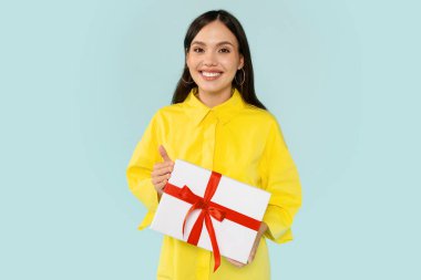 Sarı tişörtlü, elinde kırmızı fiyonklu beyaz hediye kutusu olan güzel bir kadın. Kameraya gülümsüyor, doğum günü partisine gidiyor, mavi arka planda yalnız poz veriyor.