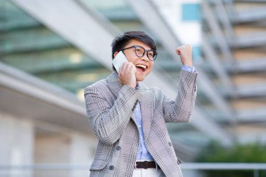 Asyalı işadamı, başarılı bir telefon görüşmesi sırasında yumruklarını sallayarak şehir caddesini modern ofis merkezinin yakınına taşıyarak kariyerinin dönüm noktasını kutluyor.