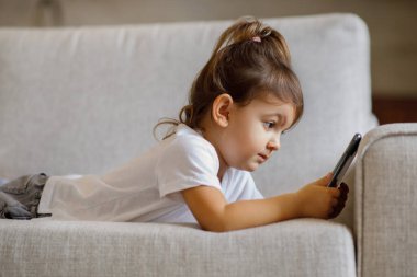 Sevimli küçük bir kız, elinde akıllı bir telefonla evde koltukta dinleniyor, küçük sevimli bir çocuk mobil oyunlar oynuyor ya da oturma odasındaki koltukta dinlenirken gelişim videoları seyrediyor,