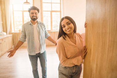 Parlak, güneşli bir mutfakta, kadın mutlu bir gülümseme ile kapıya dayanır, erkek açık açık el hareketleriyle sıcak bir hoş geldin ya da gündelik bir konuşma önerir.