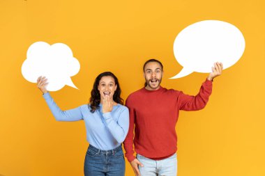 Milenyum çifti sarı stüdyo arka planında boş konuşma baloncukları tutuyor, tasarım için model alıyorlar. Çekici genç adam ve kız arkadaşı boş kelime bulutlarıyla ağızlarına dokunuyorlar.