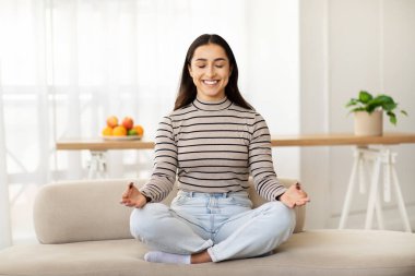 Mutlu, sakin, genç bir Arap kadın oturma odasının iç kısmında, gözleri kapalı meditasyon yapıyor. Bayan boş zamanların ve nefes egzersizlerinin keyfini çıkarın, dinlenin, rahatlayın, dinlenin ve evdeki yaşam tarzınızı değiştirin.