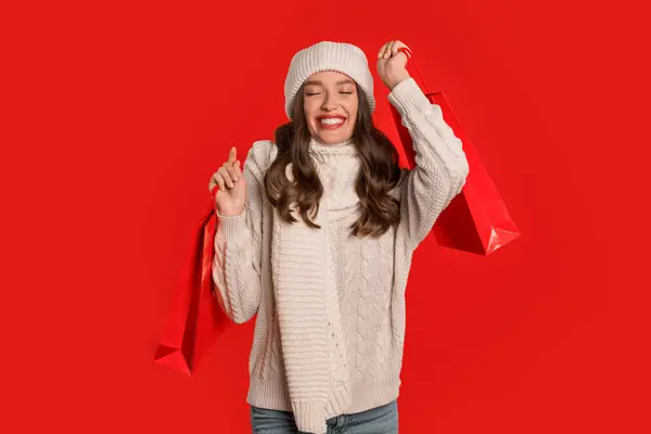 Winter holiday deals. Happy lady buyer holds shopping bags posing on red studio backdrop, celebrating start of seasonal Christmas sale. Banner with joyful shopaholic lady holding purchases