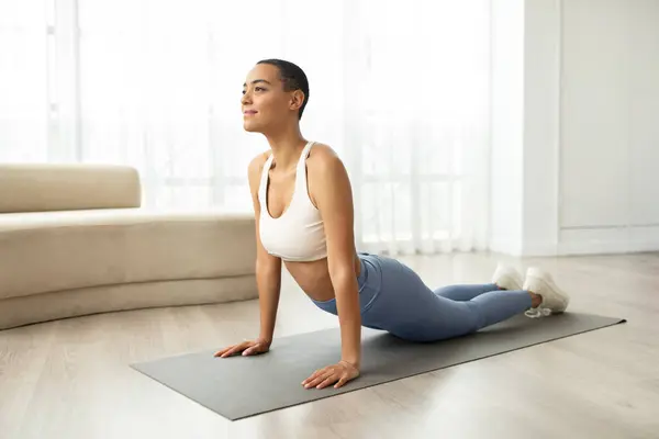 Smiling young slim latin woman in sportswear doing stretching body exercises on mat in light living room interior. Health care, weight loss and sports at home, fitness lifestyle, morning workout