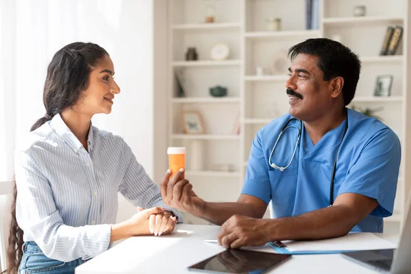 Mature Indian Doctor Man Giving Bottle With Pills To Smiling Female Patient, Male Therapist In Uniform Prescribing Medications Or Vitamins To Young Woman During Appointment In Clinic, Closeup