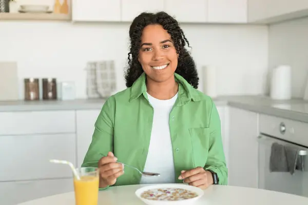 Happy black lady savoring breakfast, sitting with plate of cereals before her and glass of orange juice, enjoying meal and balanced diet in cozy kitchen at home. Nutrition and wellbeing