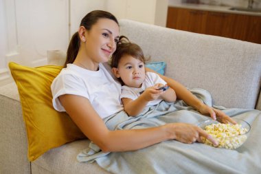 Anne-kız eğlencesi. Genç, gülümseyen bir kadın ve yürümeye başlayan çocuğu birlikte televizyon izliyorlar, sevimli küçük çocuk ve annesi koltukta uzanmış patlamış mısır yiyorlar, elinde uzaktan kumandalı çocuk