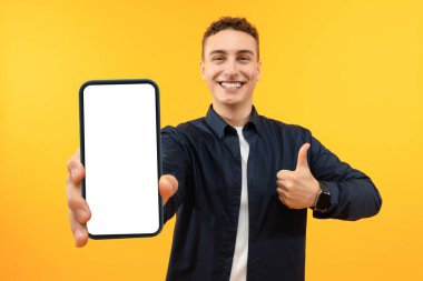 Mobil Uygulama Reklamı. Yakışıklı Heyecanlı Adam Beyaz Ekran Pozisyonu ile Akıllı Telefon Gösteriyor Sarı Stüdyo Arkaplanı Üzerinde, Kameraya Gülümsüyor. Şuna bak, cep telefonu görüntüsü.
