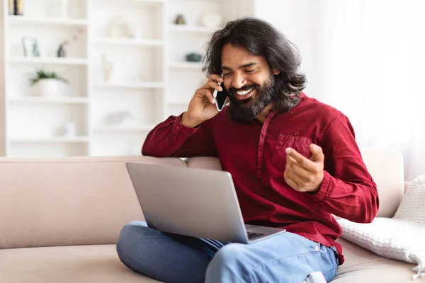 Wealthy cheerful young indian man entrepreneur working from home, sitting on couch, having phone conversation, using laptop, typing on computer keyboard, copy space. Remote business concept