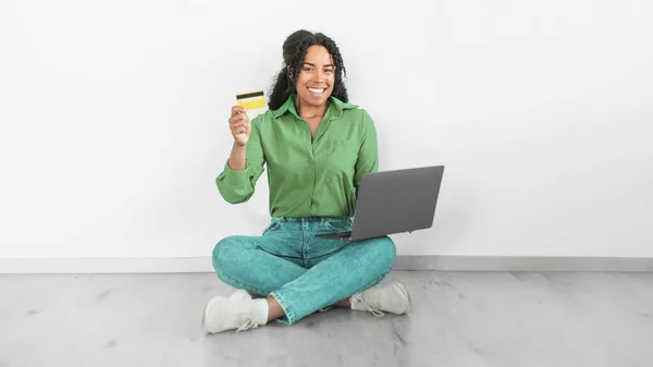 Smiling Black woman using laptop and credit card for financial transaction, symbolizing the ease of online payments and shopping, sitting on floor over white wall indoor. Ecommerce offers. Panorama