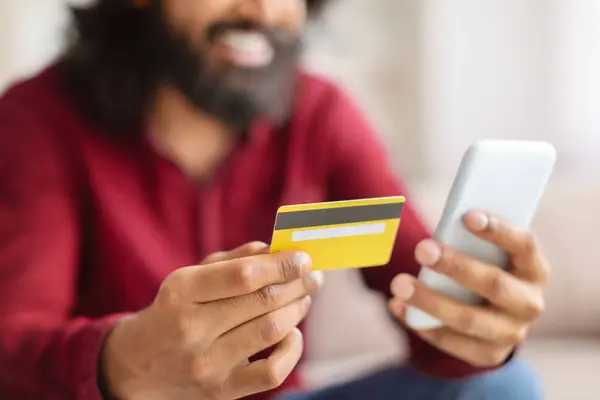 E-commerce, retail, financial app. Cropped of man in smart casual banking or shopping from home, sitting on couch in living room, using smartphone and credit card, copy space