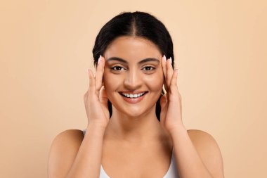 Güzellik Hizmetleri. Kameraya bakan mutlu Hintli kadın portresi kusursuz cilt teması olan, bej renkli stüdyo arka planında izole edilmiş güzel, genç ve doğulu bir kadın.