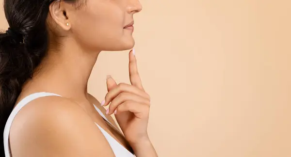 Chin Augmentation. Side view shot of unrecognizable young woman touching her face, indian female demonstrating result of plastic surgery, standing isolated on beige studio background, free space