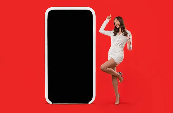 Caucasian lady in elegant dress dancing near large smartphone empty screen, holding glass of sparkling wine, while celebrating holiday event in red studio setting. Mobile app advertisement, mockup