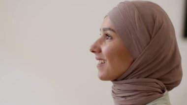 İslam güzelliği. Yan görünüşte, tesettürlü, güzel, genç bir müslüman kadının portresi. Yan tarafa bakıyor, hafifçe gülümsüyor, bej duvar arka planı, yavaş çekim, serbest alan.