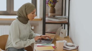 Gig ekonomi konsepti. Orta Doğulu genç kadın sekreterin bilgisayarına başörtüsü takıp klavyede yazı yazarken, ev ofisinde uzaktan çalışırken yan görüntüsü.