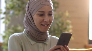 Çevrimiçi kazandı. Türbanlı haberler okuyan, akıllı telefonundan e-posta kazanan, evinde gülen mutlu müslüman bayanın yakın portresi. Başarını kutluyoruz, yavaş çekim yapıyoruz.