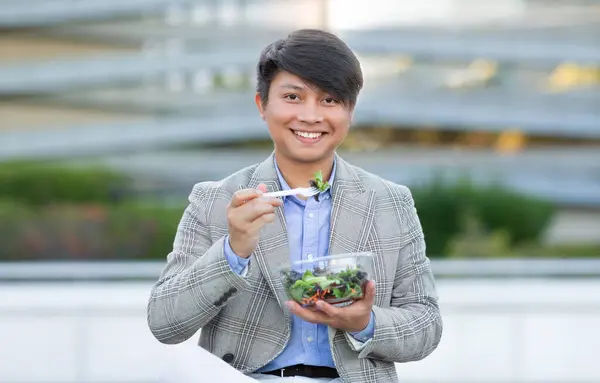 Korean entrepreneur man enjoys salad taking work break for lunch amidst urban city area outside, smiling to camera while embodying corporate lifestyle balance and healthy nutrition