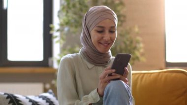 Modern online yaşam tarzı. Tesettürlü genç, kaygısız Müslüman kadın akıllı telefonuyla sörf yapıyor, kameraya bakıyor ve gülümsüyor, evinde dinleniyor, çekimin izini sürüyor, ağır çekim.