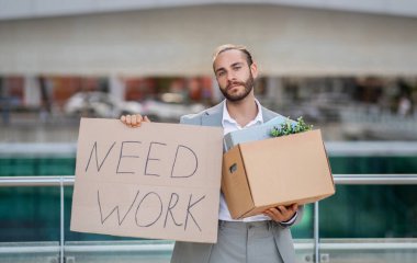 Takım elbiseli genç adam elinde karton levhayla dışarıda dururken işe ihtiyacı var, milenyum ofis çalışanı işsizlik krizi yaşıyor, işini kaybetti, işe alım sorunu yaşıyor.