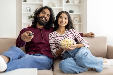 TV 'de birlikte komedi filmi izleyen mutlu Hintli eşlerin portresi, oturma odasında koltukta dinlenen doğu çifti, uzaktan kumandalı bir adamla ekranda patlamış mısır yiyen kadın.
