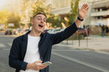 Neşeli, yakışıklı, günlük giyinmiş beyaz bir adam, şehir sokağında telefon kullanıyor, taksiyi elle durduruyor, selamlıyor, araba yakalıyor, açık havada. Ulaşım uygulaması, seyahat yaşam tarzı, acele
