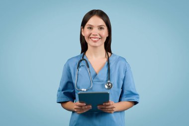 Mavi üniformalı genç Avrupalı hemşire dijital tablet tutuyor ve mavi arka planda kameraya gülümsüyor, tıbbi bilgiler sağlıyor.