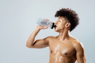 Spor yaptıktan sonra zinde genç bir adam gri stüdyo arka planında su şişesiyle poz veriyor. Mükemmel kas gücü olan tişörtsüz adam antrenman sırasında içiyor. Susuzluk ve sağlıklı zindelik.
