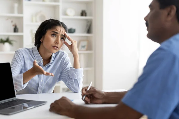 Young Indian Female Patient Sharing Health Problems With Doctor Man During Meeting In Office, Upset Sick Woman Talking To Mature Therapist, Male Physician Making Notes To Clipboard, Selective Focus