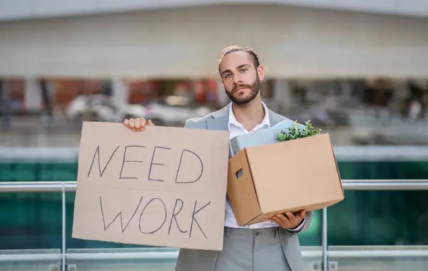 stock image Young man in suit holding cardboard sign with the text NEED WORK while standing outdoors, millennial office worker suffering unemployment crisis, lost his job, having problem with recruitment