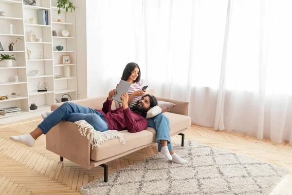 Gadget addiction, modern technologies and millennials lifestyle. High angle view of young indian spouses couple resting on sofa at home, using digital tablet and smartphone, copy space