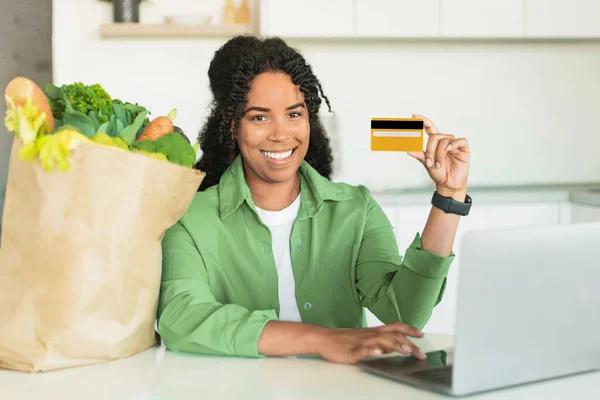 Happy young black woman with laptop computer and credit card sitting in her modern kitchen at home, advertising food products delivery and online grocery shopping offers