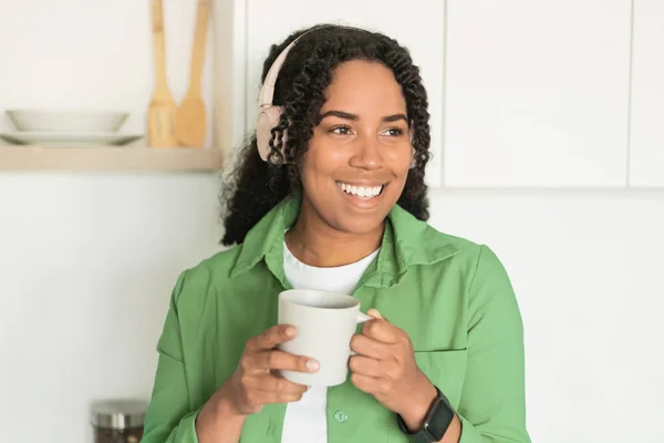 Cheerful Millennial Black Woman Drinking Coffee Enjoying Music In Headphones, Standing In Kitchen Interior, Smiling Looking Aside. Portrait Of Lady Tasting Morning Coffee And Listen To Audiobook