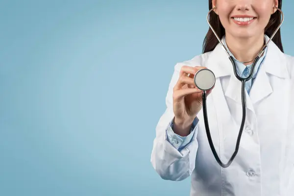 stock image Young European lady doctor in white medical uniform conducts medical checkup using stethoscope, over blue background, closeup, cropped, free space