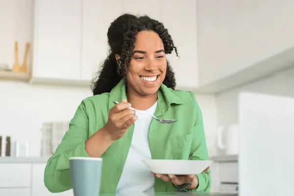 Young Black Woman Watching Videos On Laptop While Eating Cereals For Breakfast In Kitchen Interior, Happy Lady In Green Shirt Using Computer And Enjoying Tasty Oatmeal At Home