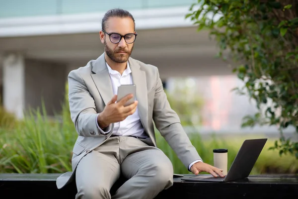 Young professional businessman in suit multitasking with smartphone and laptop while sitting on bench outdoors, handsome male entrepreneur working with mobile phone and computer outside, free space
