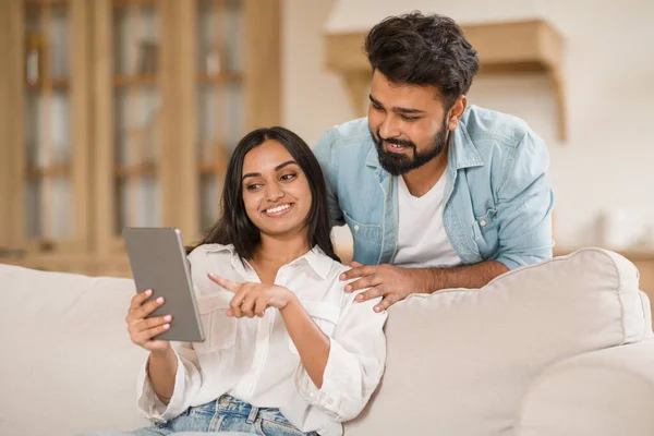 インドの女性は その画面に向かってデジタルタブレットとジェスチャーを保持しています 男は密接に傾いています 笑顔と従事の両方 快適なソファでデジタルインタラクションの瞬間を共有 — ストック写真