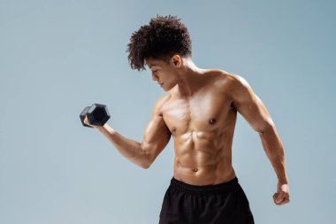 Atletik genç adam dambılları kaldırıyor, odaklanmış ve kararlı, kaslı pazıları ve gri stüdyo arka planında güçlü gövdeyi çalıştırıyor. Spor ve sağlık konsepti