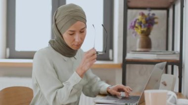 Modern iş sağlığı sorunları. Dizüstü bilgisayarda yazan tesettürlü genç müslüman bayan, gözlüğünü çıkarıyor ve burun köprüsünü ovuyor, ev ofisinde uzaktan çalışmaya devam ediyor.