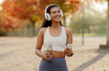 Kulaklıkla müzik dinleyen genç bir kadın, elinde su şişesi, dışarıda duruyor, sabah koşusuna ya da şehir parkında spor yapmaya hazır.