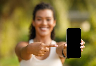 Spor kıyafetli genç bir kadın açık hava antremanı sırasında boş bir telefon ekranını işaret ediyor, internette iletişim kuruyor ve parkta uygulama modellemeleri yapıyor. Fitness teknolojisi. Seçici odak