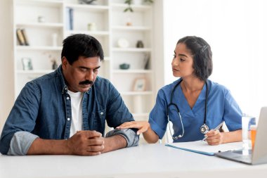 Support Concept. Young Doctor Woman Comforting Upset Indian Male Patient In Clinic, Therapist Lady Soothing Mature Man After Sharing Bad News, Touching His Hand, Expressing Empathy, Closeup clipart