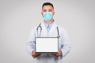 Cerrahi maske ve steteskop takan ciddi genç erkek doktor, gri zemine karşı, tıbbi danışmanlık veya sağlık uygulamaları için ideal, boş ekranlı dizüstü bilgisayar sunuyor.