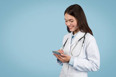 Beyaz üniformalı genç Avrupalı doktor cep telefonu kullanırken gülümsüyor, sağlık hizmetlerindeki modern teknolojiyi vurguluyor, mavi arka planda, kopyalama alanı