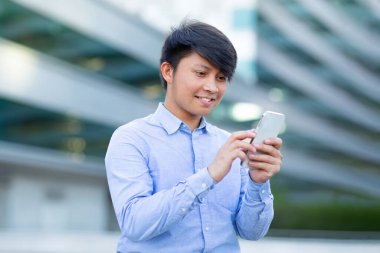 Milenyum Asyalı uzman yönetici akıllı telefonla mesajlaşıyor ve web sörfü yapıyor, şehir caddesinde modern ofis binalarıyla duruyor, teknoloji ve girişimciliğin somut bir karışımı.