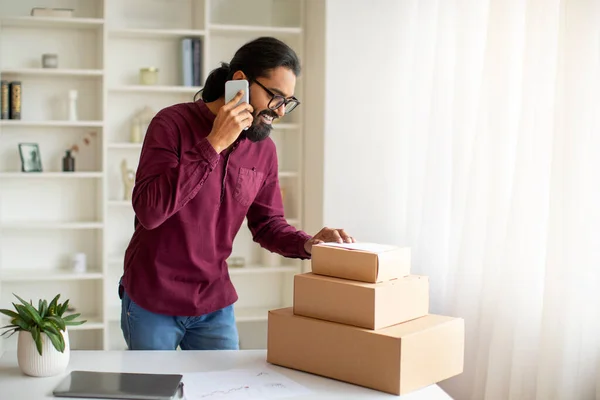 Small Business Concept. Young Indian Man Packing Boxes With Orders And Talking On Cellphone, Smiling Millennial Eastern Male Working From Home Office, Speaking With Customers By Phone, Copy Space